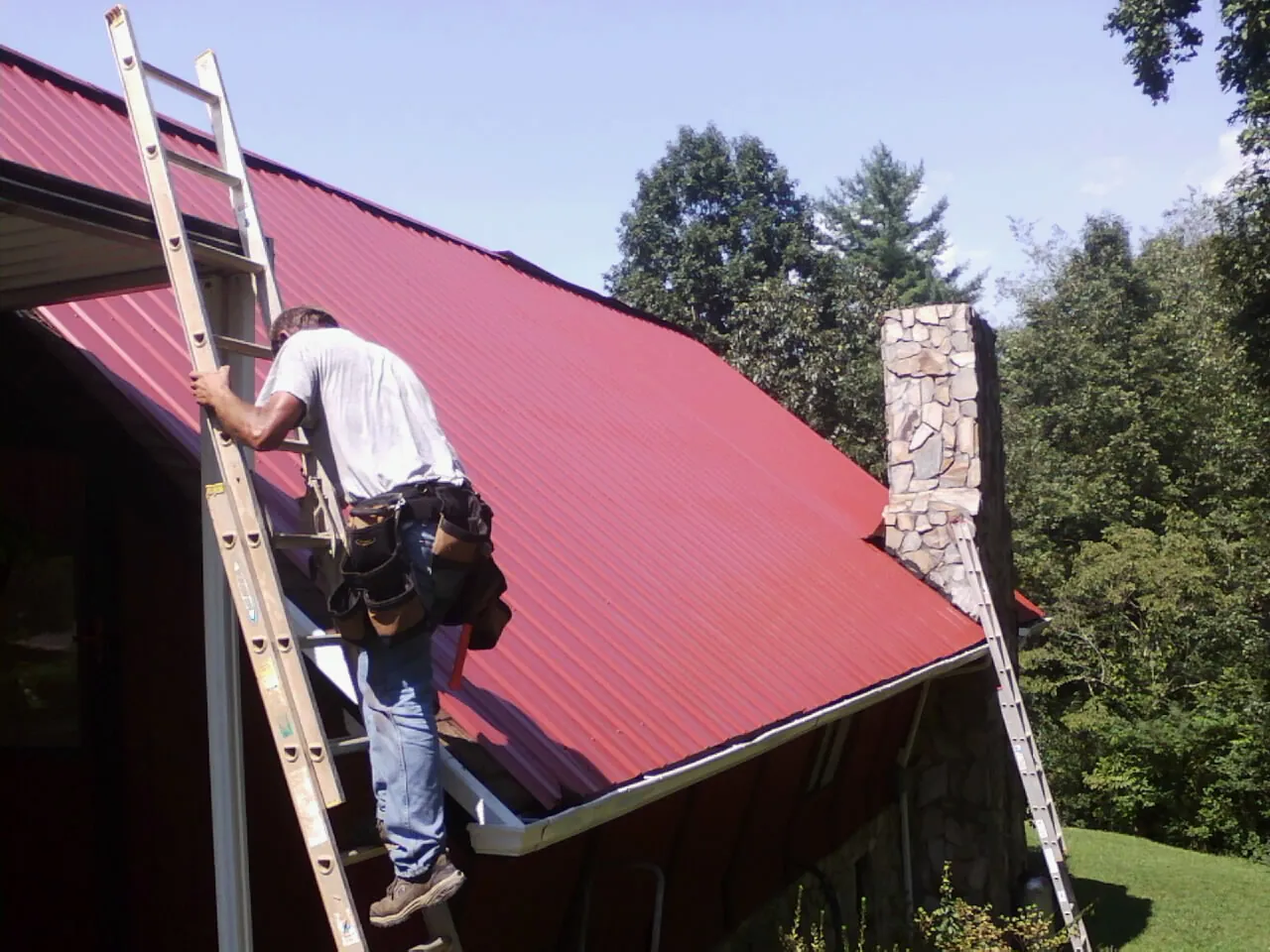 Roofer on a ladder beside a red metal roof and stone chimney — Soffit & Fascia Repair by Roofing Brothers in Kewanee